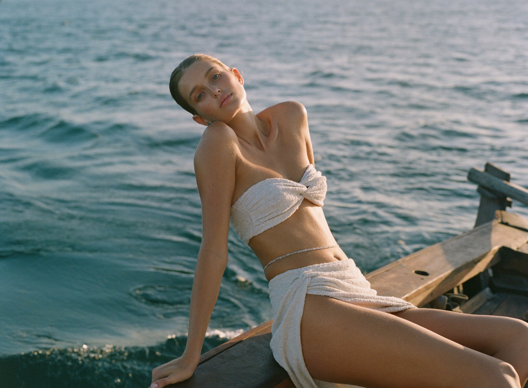 woman wearing a white bandeau top and skirt with thigh high slit, sitting on a boat with ocean background