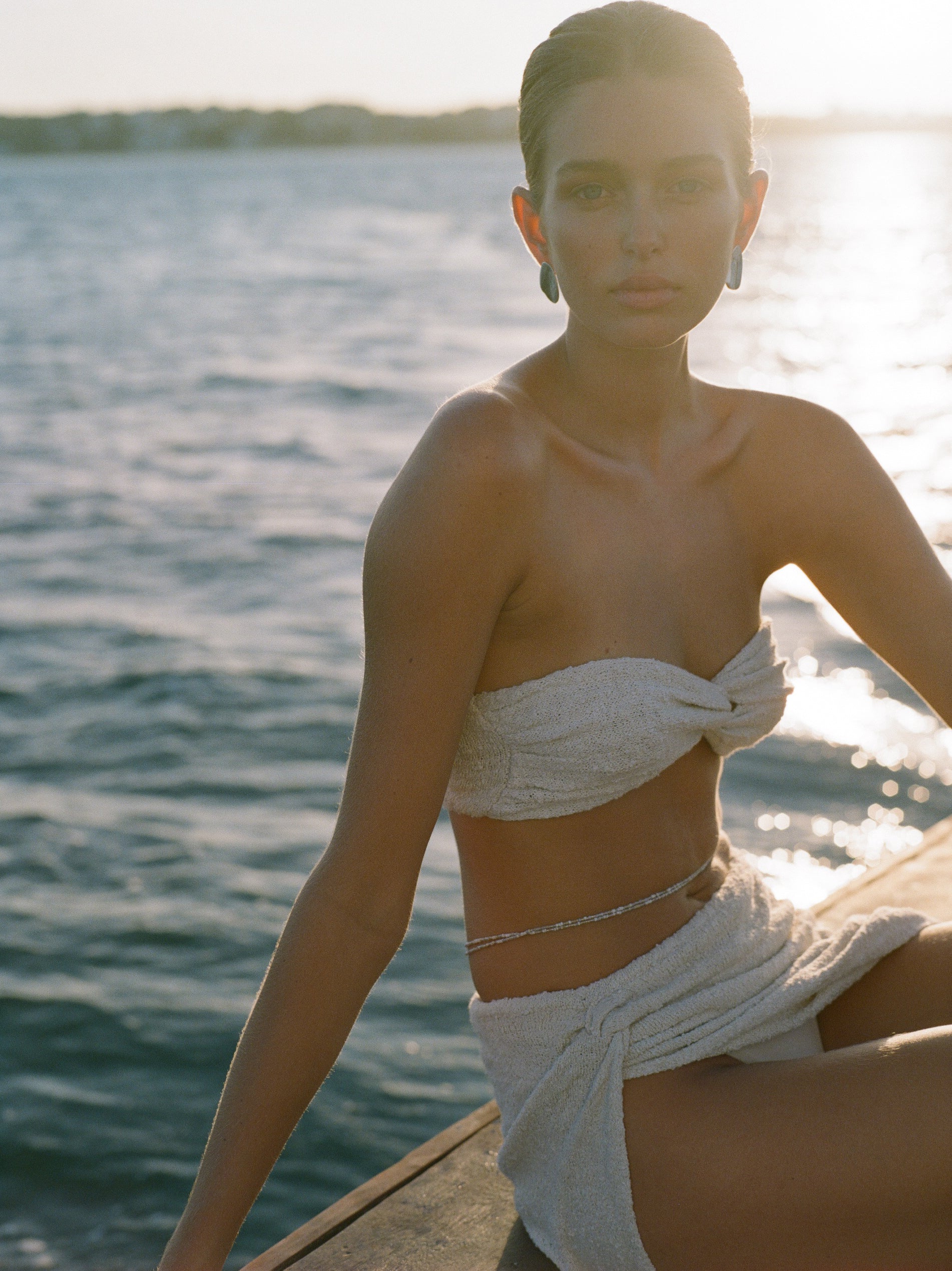 A woman sitting on a boat wearing a white bandeau-style knit top with a twist feature on the bust, paired with a high-waisted skirt.