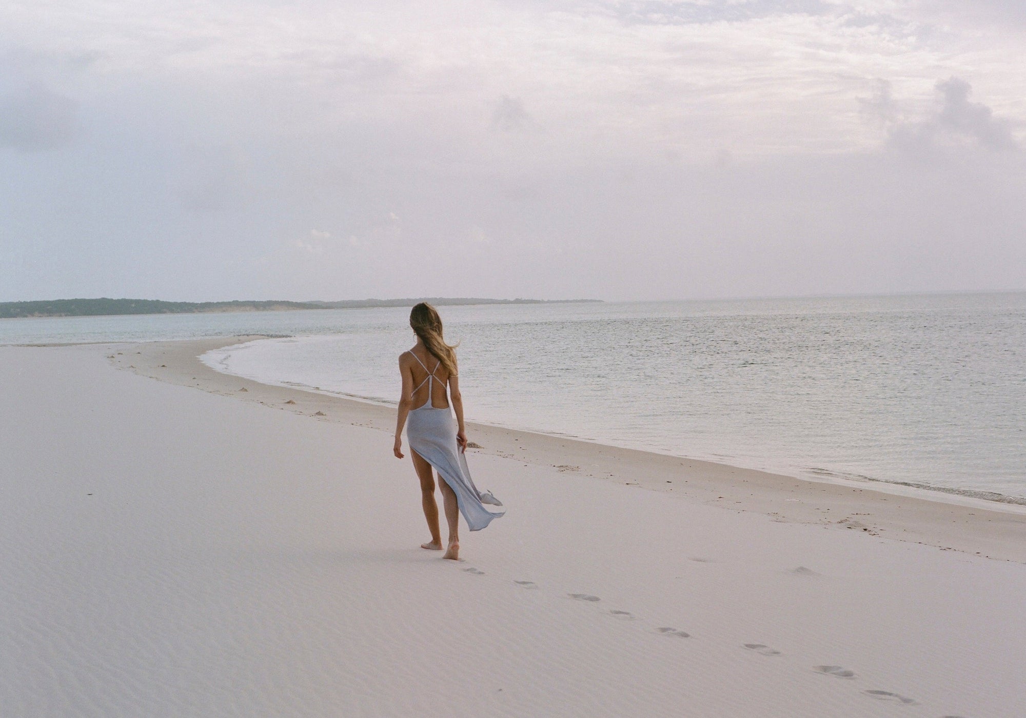 Woman in a silver blue maxi knit dress with side splits, walking on a white sand beach