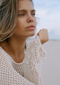 Close up of a woman wearing a shell coloured crochet inspired knit cover-up with clear tear drop beads, with a blurred background