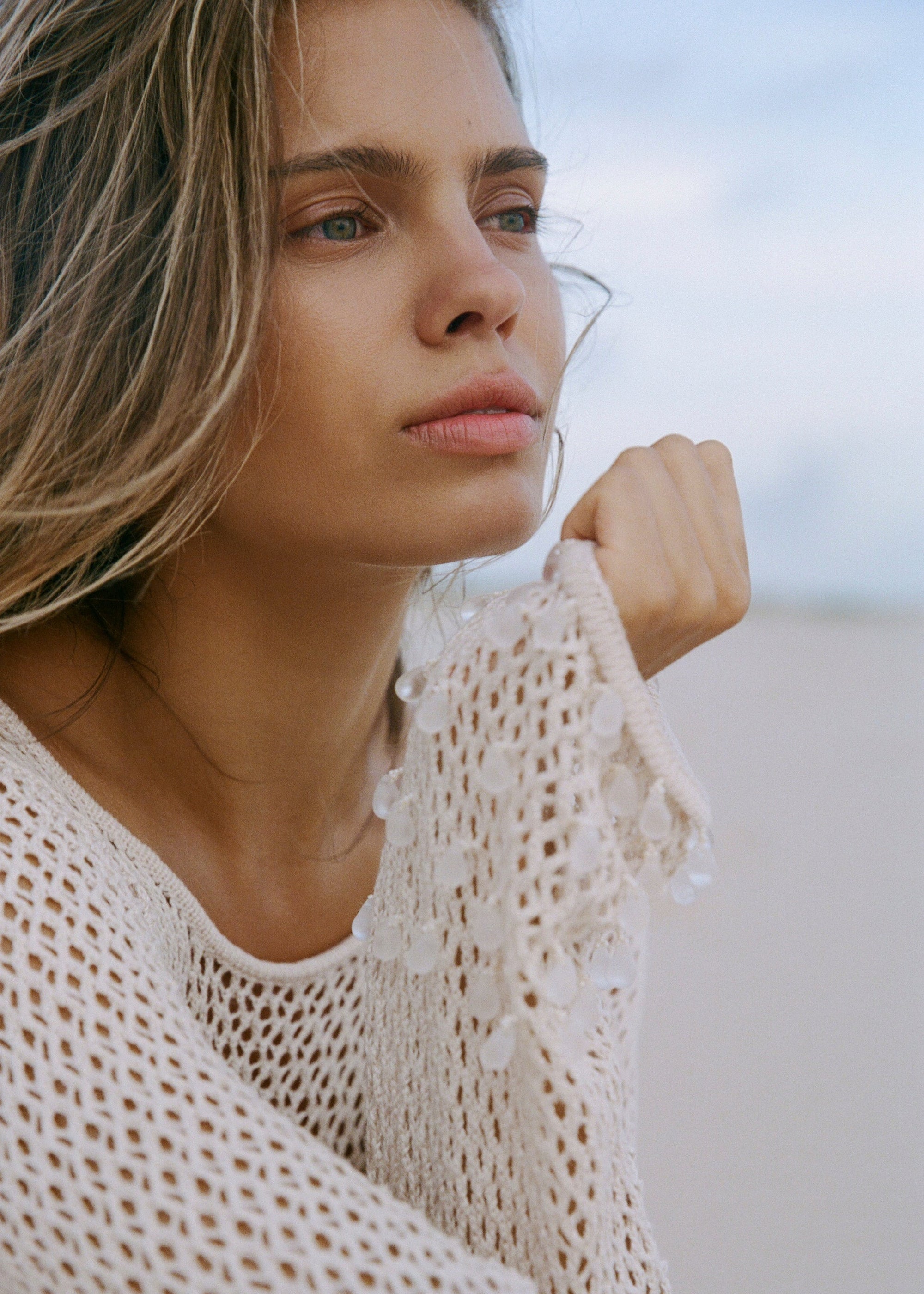 Close up of a woman wearing a shell coloured crochet inspired knit cover-up with clear tear drop beads, with a blurred background