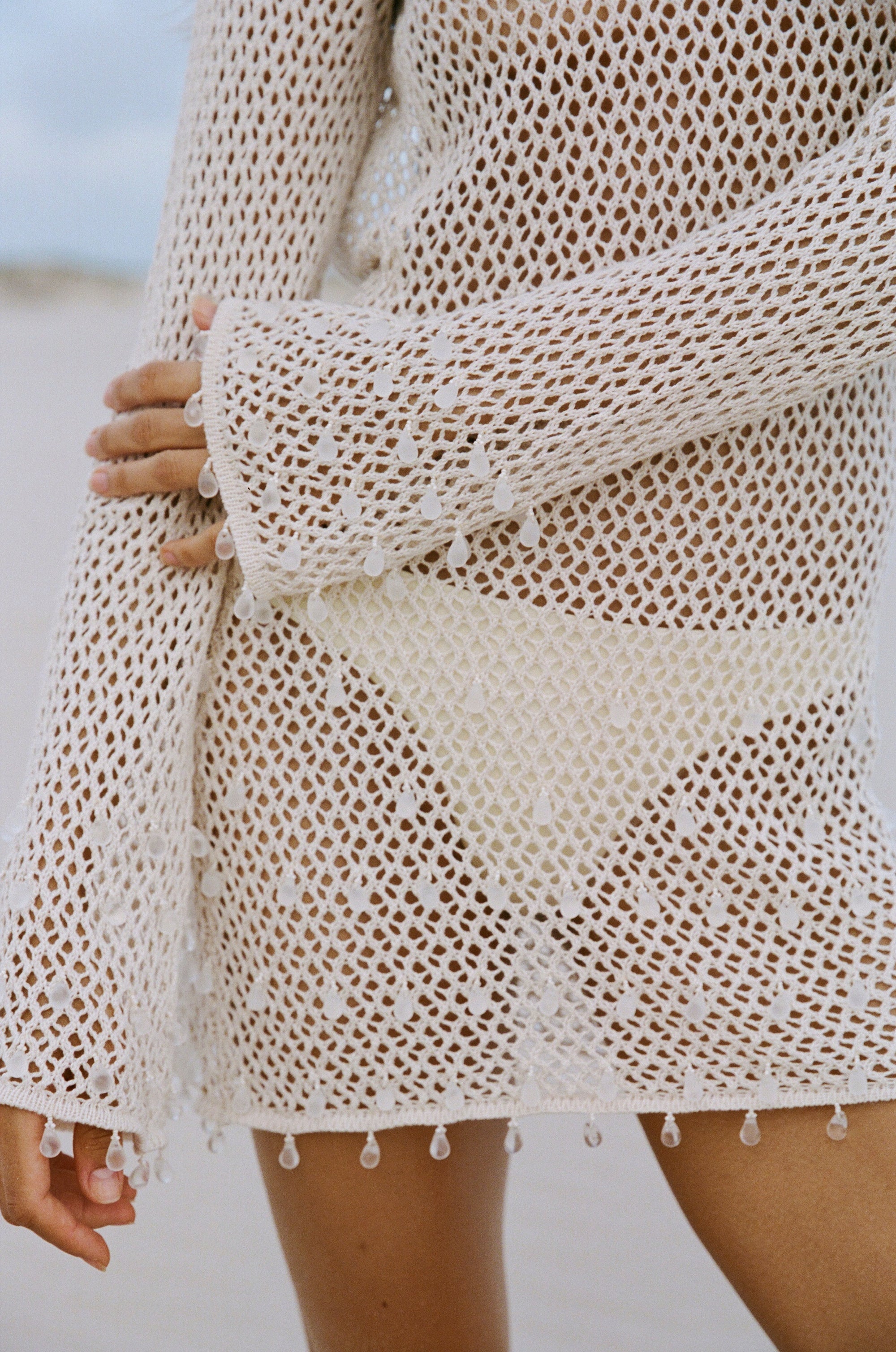 Close-up of a woman wearing a shell coloured crochet inspired knit cover-up with clear tear drop beads on a blurred background