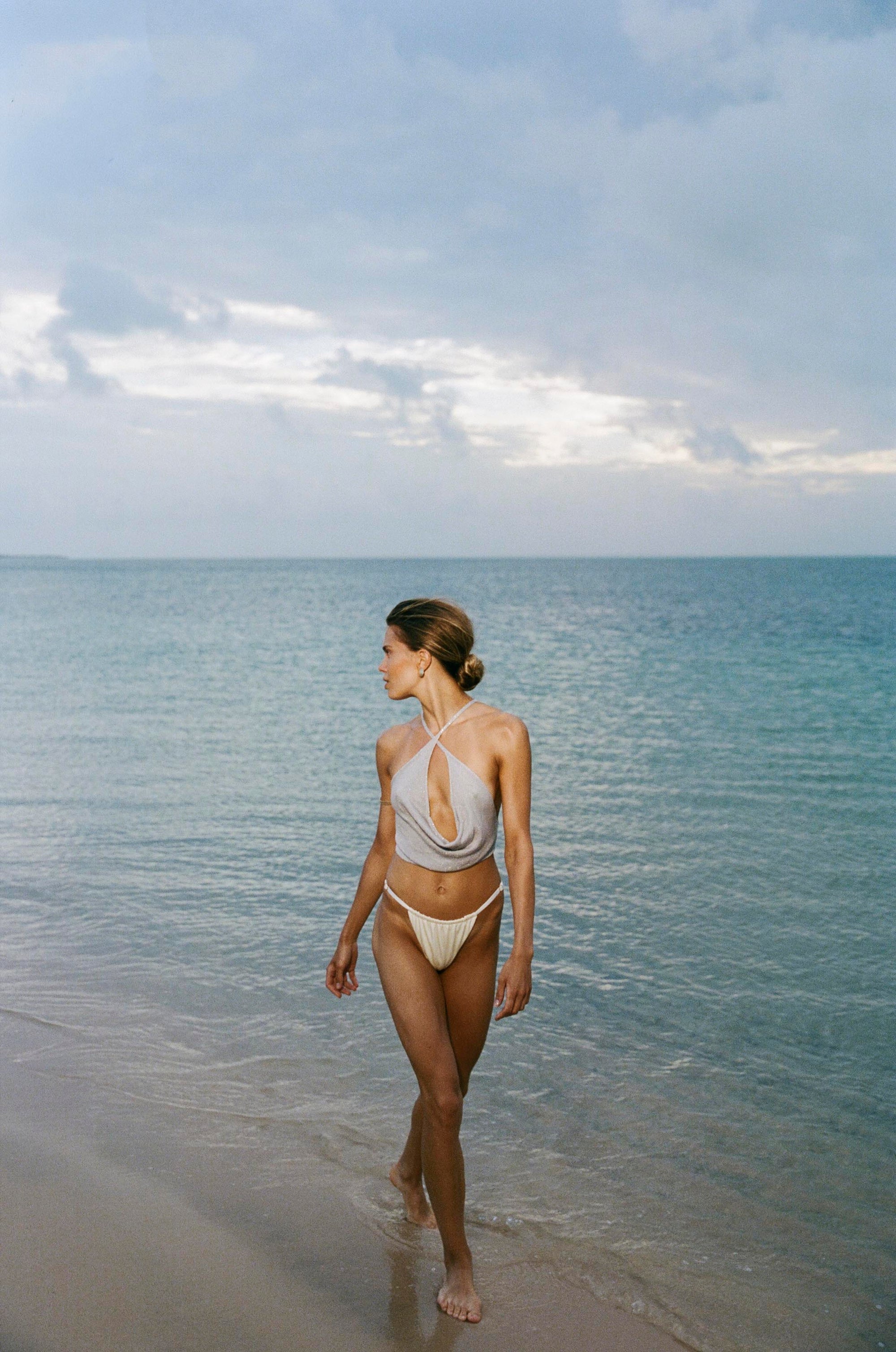 Woman in a cropped blue silver top with cowl detail and white bikini bottoms walking along a beach with a cloudy sky.