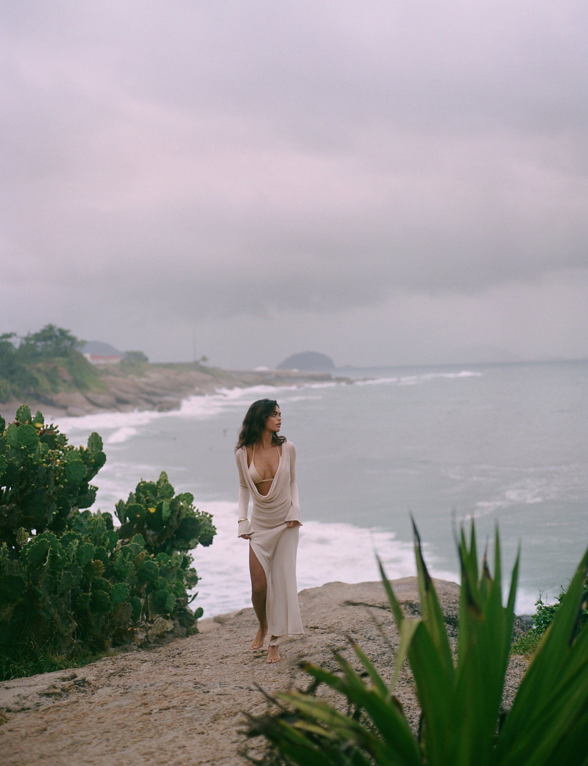 Woman in a flowing dress standing on a coastal path with ocean and overcast sky in the background