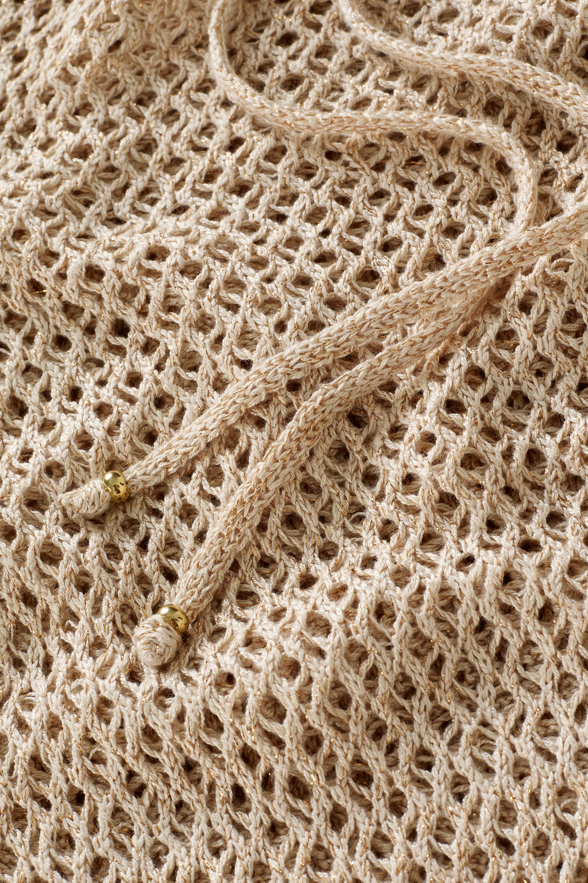 A close-up of the Isela Maxi Skirt in beige, featuring loosely woven mesh fabric and two braided drawstrings with gold bead tips.